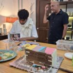 Un hombre de cabello rizado y una toalla alrededor de su cuello observa un libro en una mesa. Al lado, un hombre calvo, de pie y vestido con una camisa negra, sonríe. En la mesa hay un pastel con capas de diferentes colores y platos de comida.