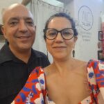 Un hombre con cabello corto y un peinado ascendido, usando una camisa negra, y una mujer con gafas grandes y un vestido rojo con estampados azules y blancos. Están de pie frente a un fondo con decoraciones artísticas y un logotipo que dice 'AVE TALLER'.