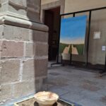 Una maceta de barro decorativa con un diseño en tonos marrones y crema, colocada sobre una base de piedra rectangular en un espacio interior. Al fondo, se ve un cuadro con una representación de un camino y un paisaje verde.