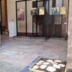 Un espacio de exposición con un panel de exhibición negro móvil que sostiene tres imágenes en un entorno arquitectónico. El suelo muestra losas de piedra y una sección decorativa con formas de madera en un marco amarillo.