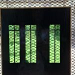 Un panel rectangular oscuro con tres secciones verticales que muestran un fondo verde. Cada sección está delimitada por líneas negras, y el panel está colgado en una malla metálica. La luz natural ilumina el área circundante.