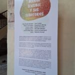 Un cartel en una estructura de soporte de madera en un espacio interior. El cartel tiene un diseño colorido en la parte superior que dice "Acrervo Sensible y sus Territorios". Incluye nombres de personas y texto explicativo sobre la exposición.