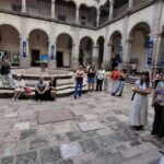 Un patio amplio con columnas y arcos en un edificio antiguo. Varias personas están de pie y sentadas en el centro, algunas se agrupan alrededor de una fuente. El cielo está parcialmente nublado, y se pueden ver carteles en las paredes del fondo.