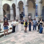 Un grupo de personas de diversas edades y estilos de vestir se reúne en un patio con columnas. Algunas personas miran al frente, otras sostienen teléfonos móviles. En el fondo, se pueden ver banners sobre cultura. El piso es de piedra.